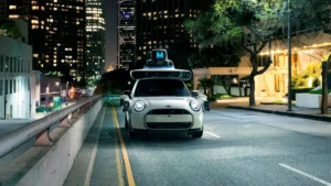 A white MINI Cooper dressed as a Waymo self-driving car drives through a quiet city street at night, featuring a mock sensor dome and “M” sign glowing on its roof. The urban skyline and streetlights frame the scene, blending playful parody with futuristic atmosphere.