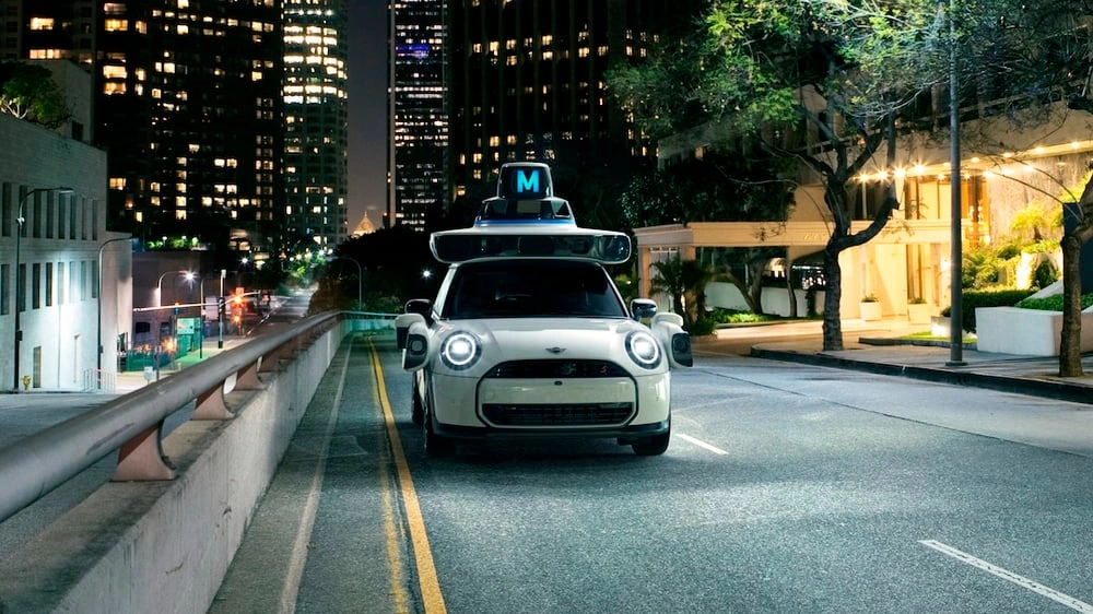 A white MINI Cooper dressed as a Waymo self-driving car drives through a quiet city street at night, featuring a mock sensor dome and “M” sign glowing on its roof. The urban skyline and streetlights frame the scene, blending playful parody with futuristic atmosphere.