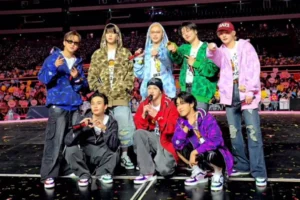 The eight members of Stray Kids pose on stage wearing their BAPE × Stray Kids Shark Hoodies in distinct camouflage colours — blue, green, purple, pink, red, brown, and sky-blue — with matching BAPE STA sneakers. The group performs in front of a brightly lit arena crowd, highlighting the fusion of K-pop energy and Japanese streetwear aesthetics.