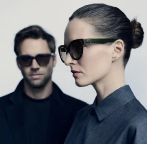 Close-up portrait of a woman and man wearing Robert Marc NYC eyewear. The woman, in front, wears oversized green acetate frames and a sleek grey shirt, her hair styled in a minimalist bun. Behind her, the man wears dark rectangular sunglasses with a tailored navy coat. The image captures Robert Marc NYC’s Fall 2025 mood — modern refinement, quiet confidence, and timeless New York elegance.