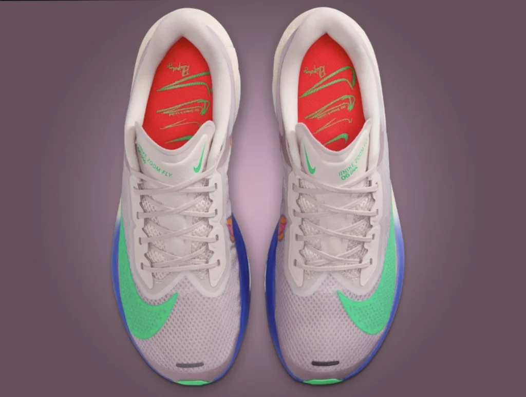 Top-down view of the Nike Zoom Fly “No Human Is Limited” sneakers designed with Eliud Kipchoge’s influence. The shoes feature pale lilac mesh uppers, bright green Swooshes, and vivid red insoles signed with Kipchoge’s name and motivational graphics. The midsoles blend deep blue and orange tones, with “No Human Is Limited” subtly marked on the side, all set against a mauve background emphasizing the shoes’ bold, aerodynamic silhouette.