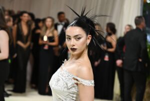 Charli XCX poses on the red carpet wearing an off-shoulder white gown with distressed detailing and bold dark lipstick, her hair styled in an avant-garde spiked updo, surrounded by attendees in formal evening wear at a gala event.
