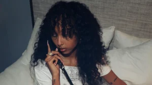 Singer Girlfriend sits on a bed holding a black corded phone, gazing thoughtfully to the side. She wears a white printed tee, gold rings, and layered necklaces, with voluminous curly hair framing her face. The soft lighting and intimate setting evoke a moody, late-night aesthetic that complements her pop persona.