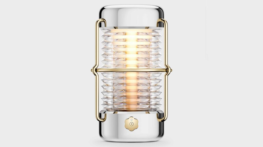 Front view of the Balmuda “Sailing Lantern” designed by Jony Ive, featuring a ribbed glass cylinder encased in polished aluminum and gold-toned brass, emitting a warm ambient glow.
