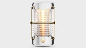 Front view of the Balmuda “Sailing Lantern” designed by Jony Ive, featuring a ribbed glass cylinder encased in polished aluminum and gold-toned brass, emitting a warm ambient glow.
