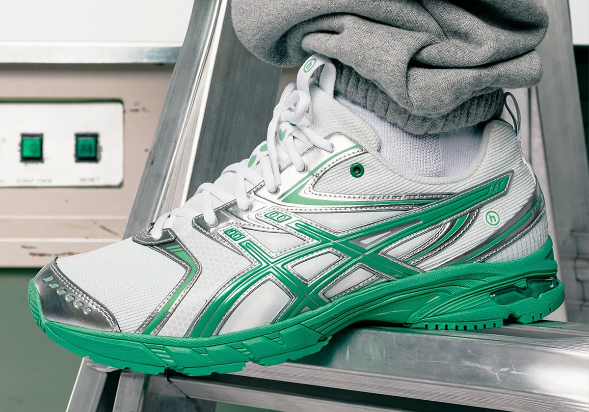 Close-up of the HIDDEN x ASICS GEL-DS Trainer 14 sneaker in white mesh with metallic chrome overlays and bright green detailing, worn with grey sweatpants, photographed on a metal ladder in a minimalist studio setting.