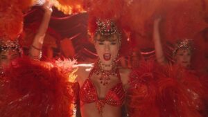 Taylor Swift performing in a vibrant showgirl-inspired red costume adorned with sequins, beads, and feathered accessories, surrounded by dancers in matching outfits under warm stage lighting — a scene evoking classic cabaret glamour from The Fate of Ophelia music video.
