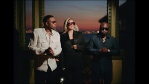 Nas, Jorja Smith, and Havoc of Mobb Deep stand side by side on a bridge at sunset, dressed in sleek black and white outfits with dark sunglasses. The warm glow of dusk and distant water create a cinematic, reflective atmosphere, blending elegance with urban cool.
