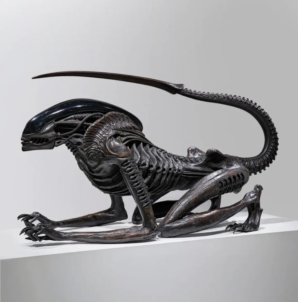 Life-size Necronom sculpture by H.R. Giger, created for Alien III, depicting a crouched biomechanical creature with an elongated, ridged tail and glossy black exoskeletal head, blending anatomical and mechanical details; displayed on a white plinth at Mai 36 Galerie during H.R. Giger Paris exhibition, 2025.