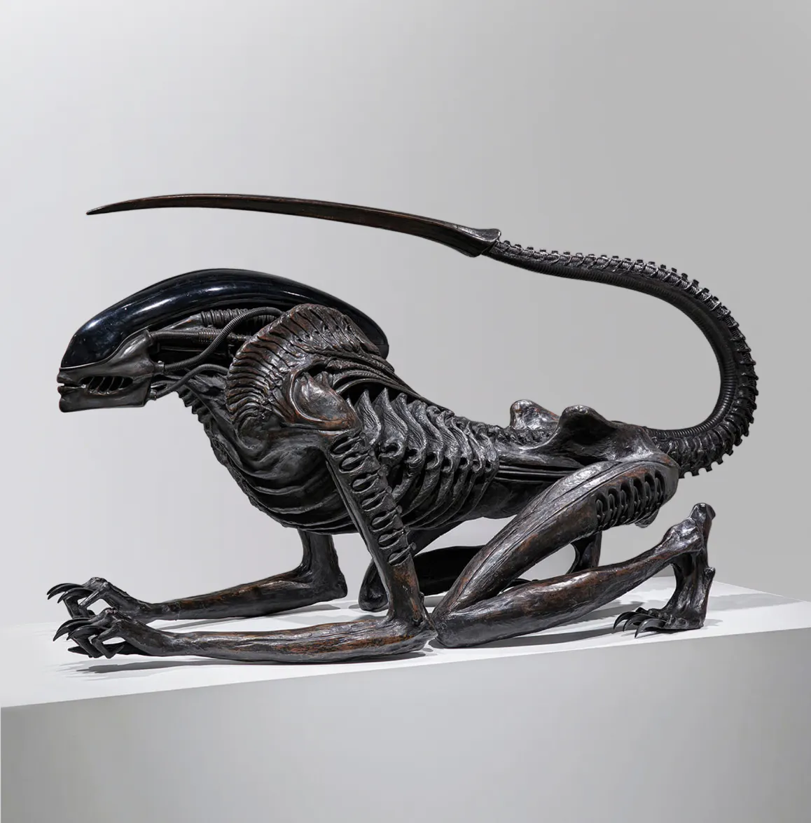 Life-size Necronom sculpture by H.R. Giger, created for Alien III, depicting a crouched biomechanical creature with an elongated, ridged tail and glossy black exoskeletal head, blending anatomical and mechanical details; displayed on a white plinth at Mai 36 Galerie during H.R. Giger Paris exhibition, 2025.