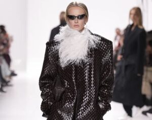 Model walking the runway in Bottega Veneta’s Spring/Summer 2026 show wearing a dark woven leather oversized coat with a dramatic white feather collar, paired with sleek black sunglasses and a clutch.