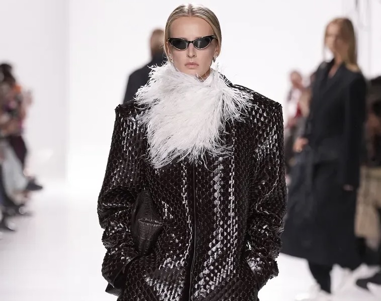 Model walking the runway in Bottega Veneta’s Spring/Summer 2026 show wearing a dark woven leather oversized coat with a dramatic white feather collar, paired with sleek black sunglasses and a clutch.