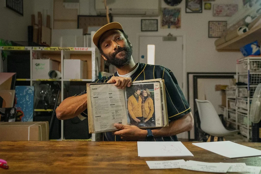 James: wearing a yellow cap and striped shirt sits at a wooden desk, holding open a vintage knitting pattern book that shows a man and woman in matching yellow cardigans, inside an art studio filled with boxes, shelves, and framed works.