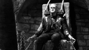 A black-and-white still from Son of Frankenstein (1939) showing the Frankenstein Monster strapped to a large stone chair inside a dark laboratory. Heavy metal restraints hold his head and arms in place, while chains hang from the surrounding stone archway. The Monster’s lifeless expression and the harsh lighting emphasize the film’s gothic tension and tragic undertones.