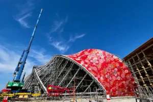The Hosoo pavilion under construction at Expo 2025 Osaka, showing a vast red curved structure wrapped in floral Nishijin brocade fabric with cranes and scaffolding beneath a clear blue sky.