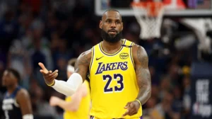 LeBron James, wearing a yellow Los Angeles Lakers jersey with the number 23, gestures with his hand during an NBA game, looking focused with the crowd blurred in the background.