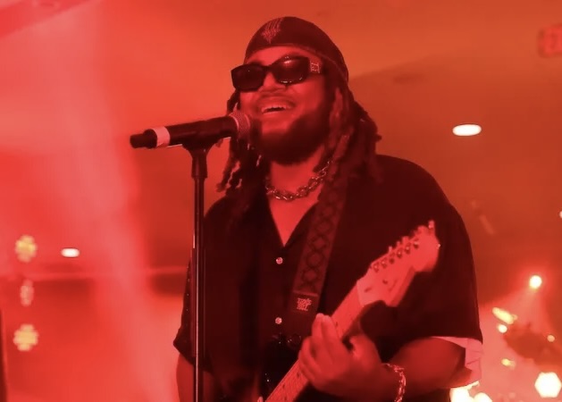 Thomas Leon performing live under red stage lights, smiling while playing an electric guitar and singing into a microphone and wearing sunglasses.