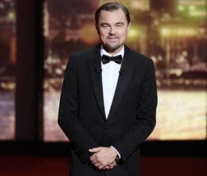 Leonardo DiCaprio stands on stage at the 2025 Cannes Film Festival in a classic black tuxedo and bow tie, hands clasped, smiling calmly under warm lights, reflecting the timeless poise and understated showmanship that define his public presence.