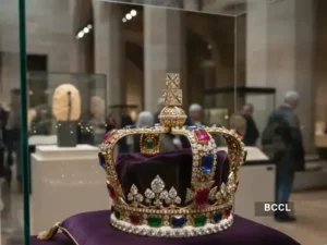 Crown of Empress Eugénie displayed inside a glass case at the Louvre Museum in Paris. The ornate gold crown is set with large emeralds, rubies, and diamonds, resting on a purple velvet cushion under museum lighting, with visitors blurred in the background.