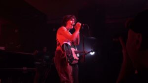 King Princess on stage under red lighting, holding a microphone and playing an electric guitar.