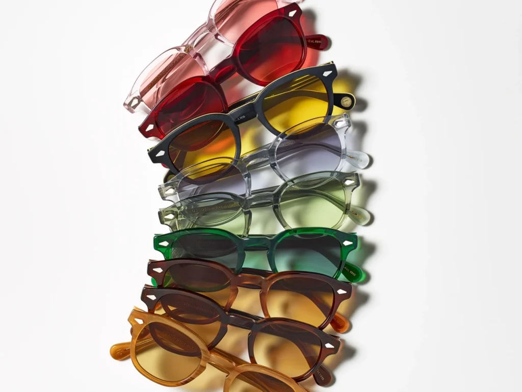 A neatly stacked arrangement of MOSCOT Lemtosh sunglasses displayed diagonally on a white surface, showcasing translucent acetate frames in a spectrum of colors — pink, red, yellow, clear gray, green, brown, and amber — each fitted with matching tinted lenses that highlight the brand’s craftsmanship and playful tonal variety.