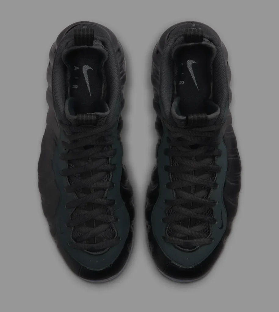 Top-down view of the Nike Air Foamposite One “Carbon Fiber” sneakers showing the symmetrical black laces, mesh tongues with pull tabs, and subtle silver Nike Swoosh logos on the insoles, set against a grey background emphasizing the sculpted foam contours and snug boot-like fit.