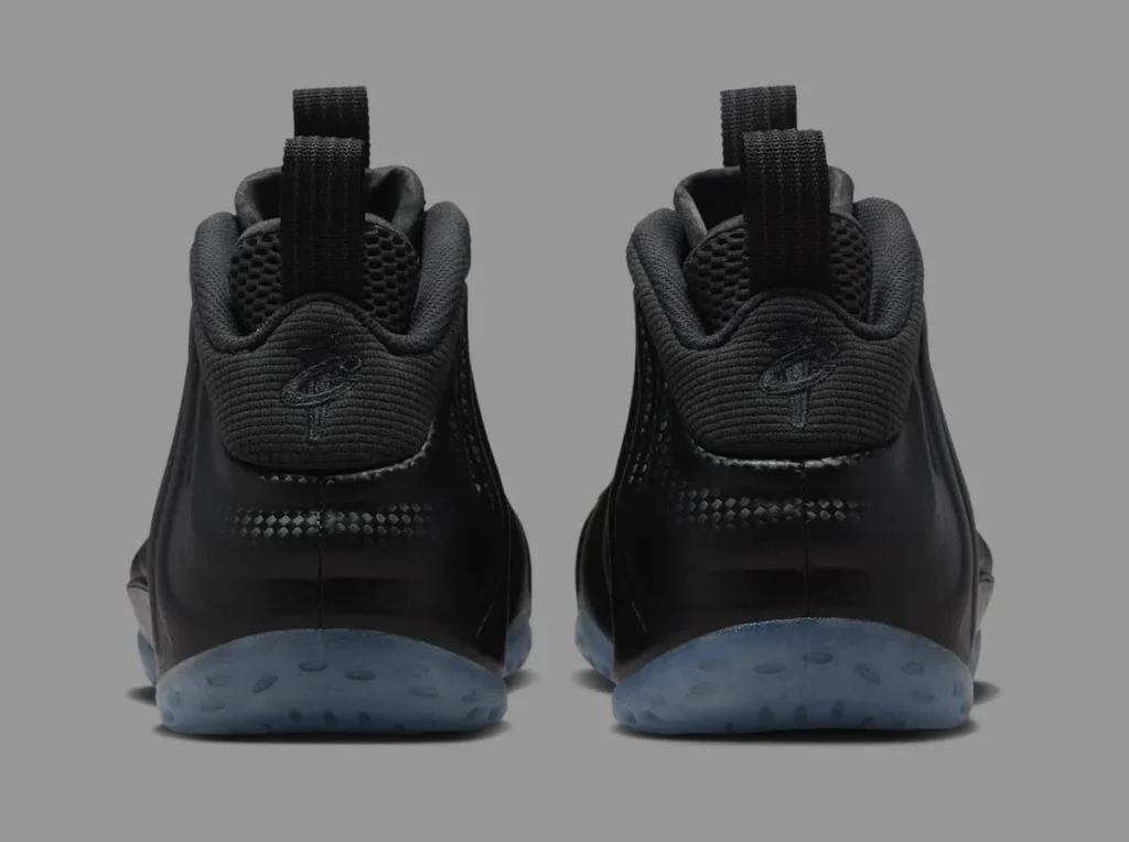 Rear view of the Nike Air Foamposite One “Carbon Fiber” sneakers showing black ribbed heels with embroidered Penny Hardaway “1 Cent” logos, mesh pull tabs, and translucent icy blue outsoles — photographed against a grey background to highlight the shoe’s molded carbon-textured shell and sleek, symmetrical build.