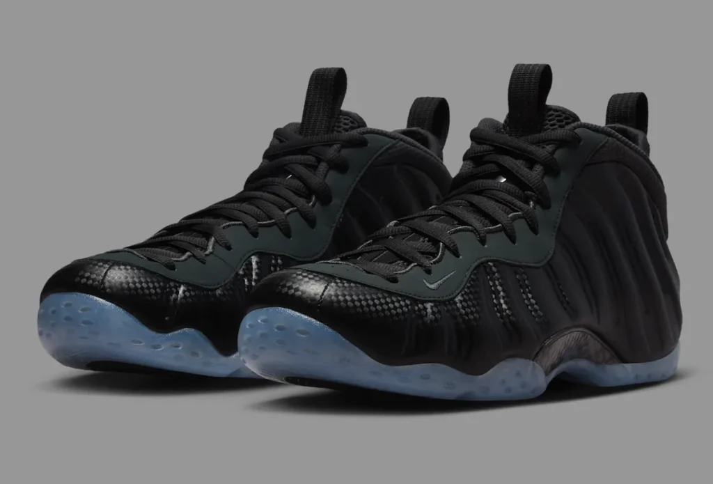 Nike Air Foamposite One “Carbon Fiber” shoes featuring a black molded upper with carbon weave detailing, matte black eyestays, mesh tongue, and translucent icy blue outsole — displayed against a neutral grey background to emphasize the shoe’s futuristic sculpted silhouette and reflective texture.