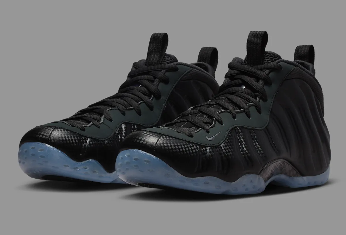 Nike Air Foamposite One “Carbon Fiber” shoes featuring a black molded upper with carbon weave detailing, matte black eyestays, mesh tongue, and translucent icy blue outsole — displayed against a neutral grey background to emphasize the shoe’s futuristic sculpted silhouette and reflective texture.
