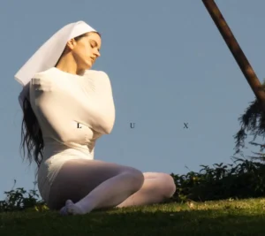 Rosalía kneeling on grass under clear daylight, dressed in a white fitted gown and veil, arms crossed in serene posture, embodying minimalist religious imagery for her upcoming album LUX.