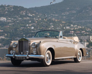 1962 Rolls-Royce Silver Cloud II Drophead Coupé Adaptation by H.J. Mulliner parked against a mountainous Mediterranean backdrop, finished in pale metallic gold with ivory convertible top, representing the final coachbuilt example of its kind.