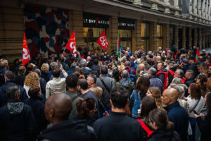 "Crowd of protesters gathered outside Le BHV Marais in Paris holding red union flags, demonstrating against Shein’s upcoming boutique and fast-fashion expansion.”
