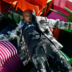 Virgil Abloh reclines on a stack of brightly colored plastic chairs, wearing a black Supreme T-shirt, splattered black pants, and a patterned jacket. The photograph captures his effortless blend of art, streetwear, and design, reflecting the creative energy that defined his multidisciplinary vision.