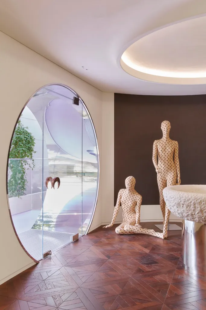 Interior of Marine Serre’s Hannam House in Seoul featuring two crescent-moon–patterned mannequins beside a circular glass entrance door, a sculpted stone basin on a metal pedestal, and warm parquet wood flooring under soft halo lighting.