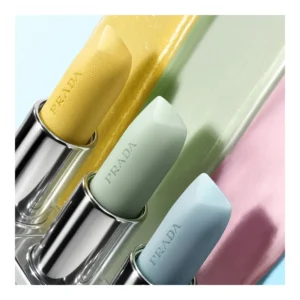 Close-up image of three Prada lip balms in sleek metallic cases, displayed in soft pastel hues of yellow, mint green, and powder blue. Each bullet is embossed with the PRADA logo and set against smooth swatches of balm textures in yellow, green, and pink, highlighting the luxurious, creamy consistency and modern color palette of the Prada Balm collection.