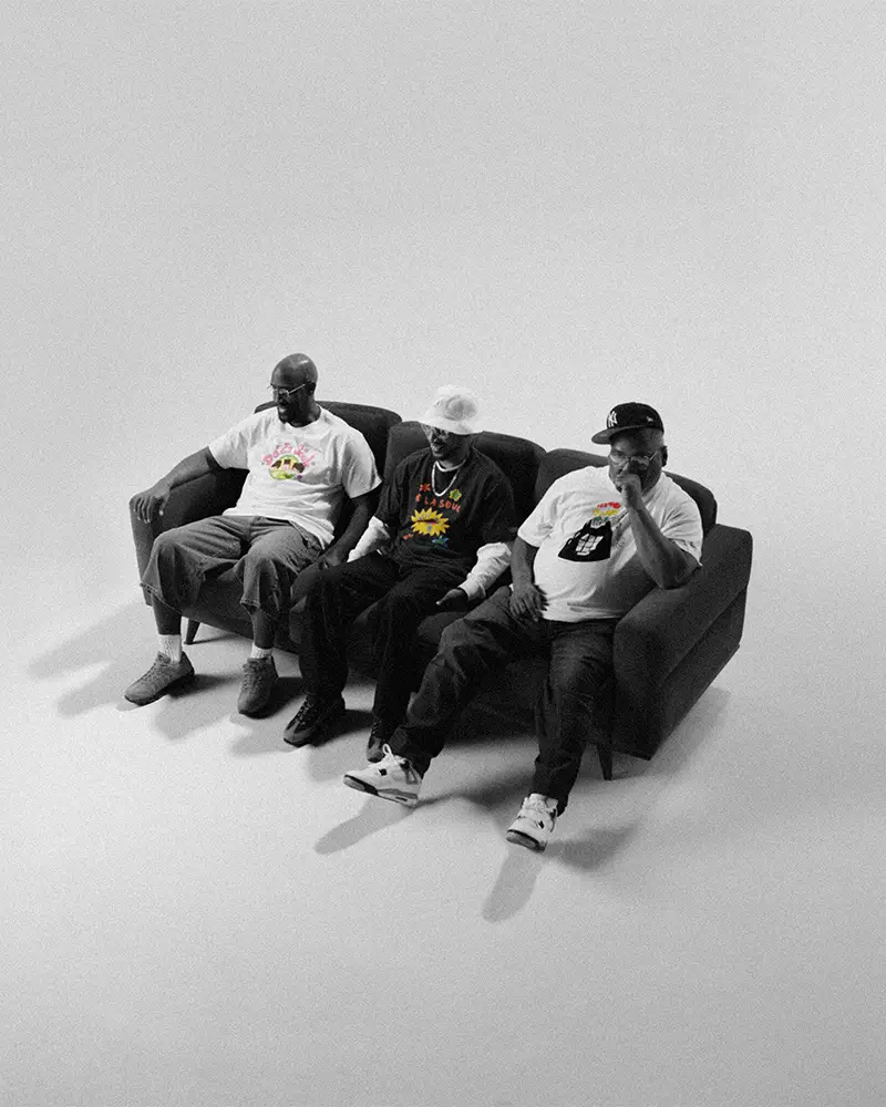 Black-and-white photo of three seated men wearing graphic De La Soul x Levi’s T-shirts, sitting on a sofa in a minimal studio space