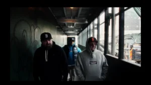 Slaine, Statik Selektah, and ILL BILL walk through a narrow, dimly lit city passageway lined with graffiti and metal fencing, dressed in hoodies and baseball caps representing Boston and New York. The scene captures a gritty, cinematic mood that echoes the raw tone of their “Cancel Culture” collaboration.