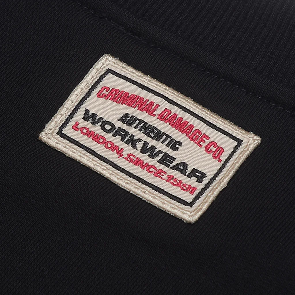 Close-up of the stitched Criminal Damage workwear patch on the back neckline of the sweatshirt, featuring red and black typography on an off-white canvas label against a black fabric background