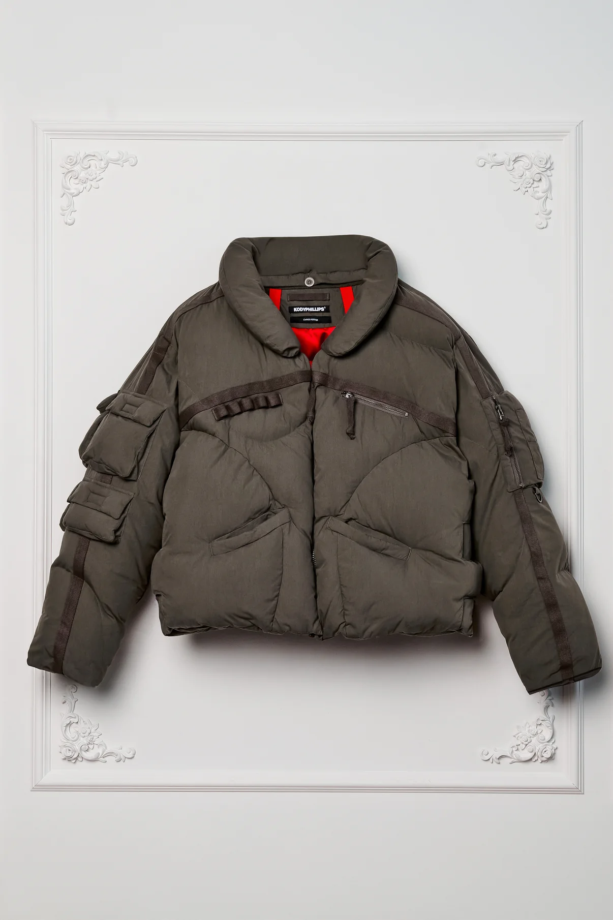 Front view of the Kody Phillips Cargo Puffer in Sage, showcasing its structured silhouette, multiple cargo pockets on the sleeves, and heavy-duty webbing details, contrasted by a glimpse of its vivid red interior lining