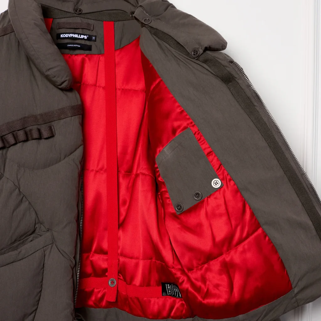 Interior view of the Kody Phillips Cargo Puffer in Sage showing the striking red satin lining, internal pocket with button closure, and backpack-style straps integrated into the design