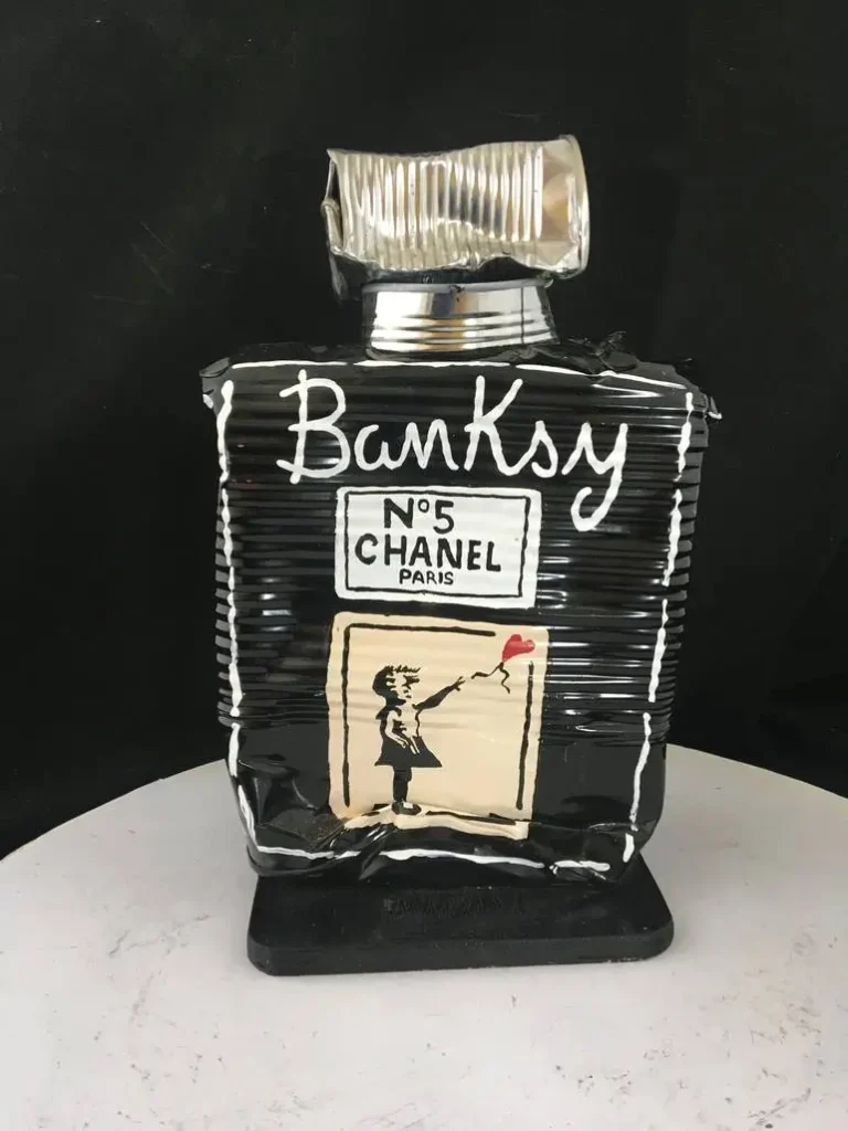 A black, crushed-metal sculpture shaped like a Chanel No. 5 perfume bottle, painted with the word “Banksy” in white and featuring a stencil-style image of a girl releasing a red heart-shaped balloon