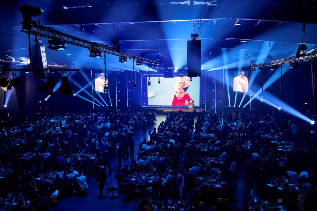View of the 2025 UK Music Video Awards ceremony inside Magazine London, showing a packed audience seated at round tables beneath blue stage lighting as a large screen displays a performance still and the presenter on stage.