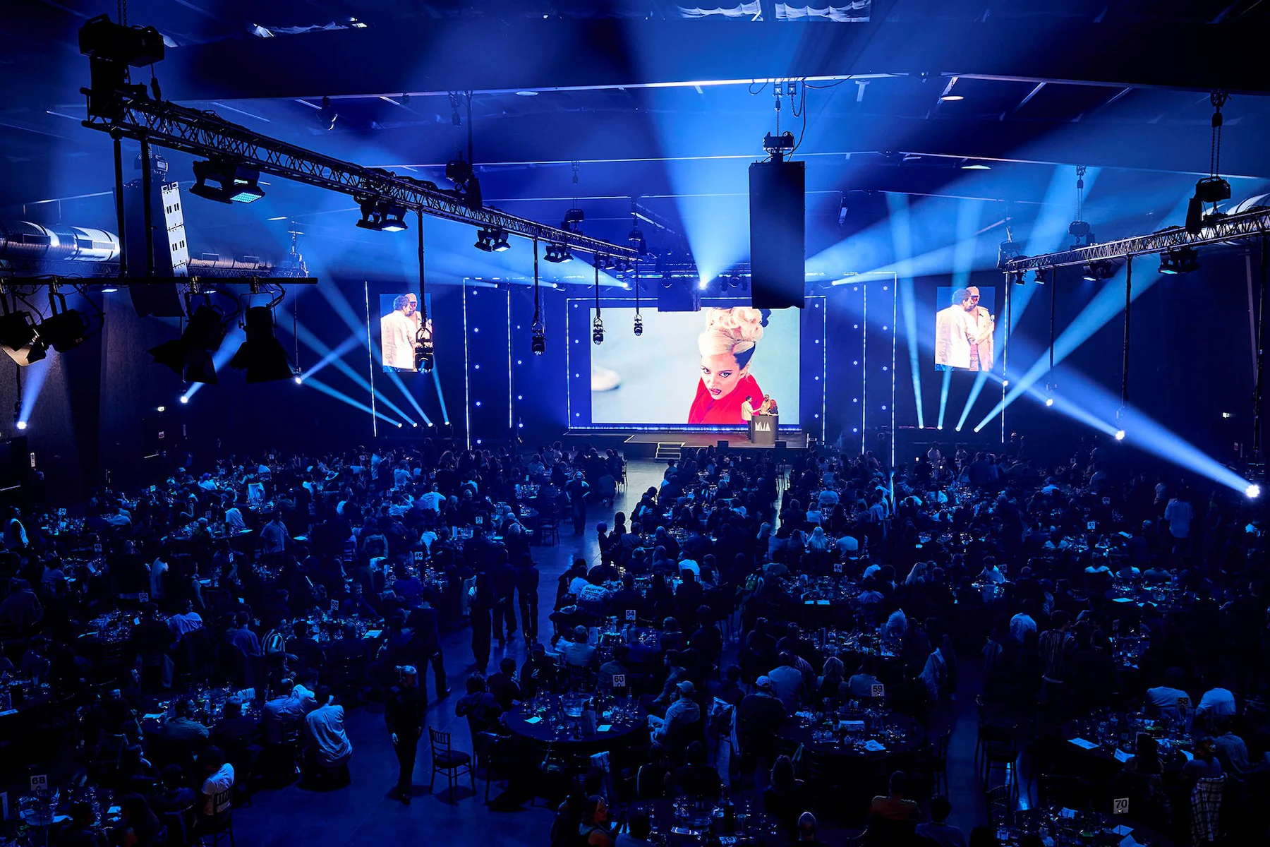 View of the 2025 UK Music Video Awards ceremony inside Magazine London, showing a packed audience seated at round tables beneath blue stage lighting as a large screen displays a performance still and the presenter on stage.