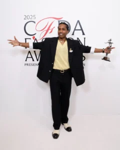 A$AP Rocky poses with his CFDA Fashion Icon Award at the 2025 CFDA Fashion Awards in New York, wearing a black tailored suit over a pale yellow knit shirt, accessorized with layered gold jewelry and two-tone shoes, extending his arms in celebration