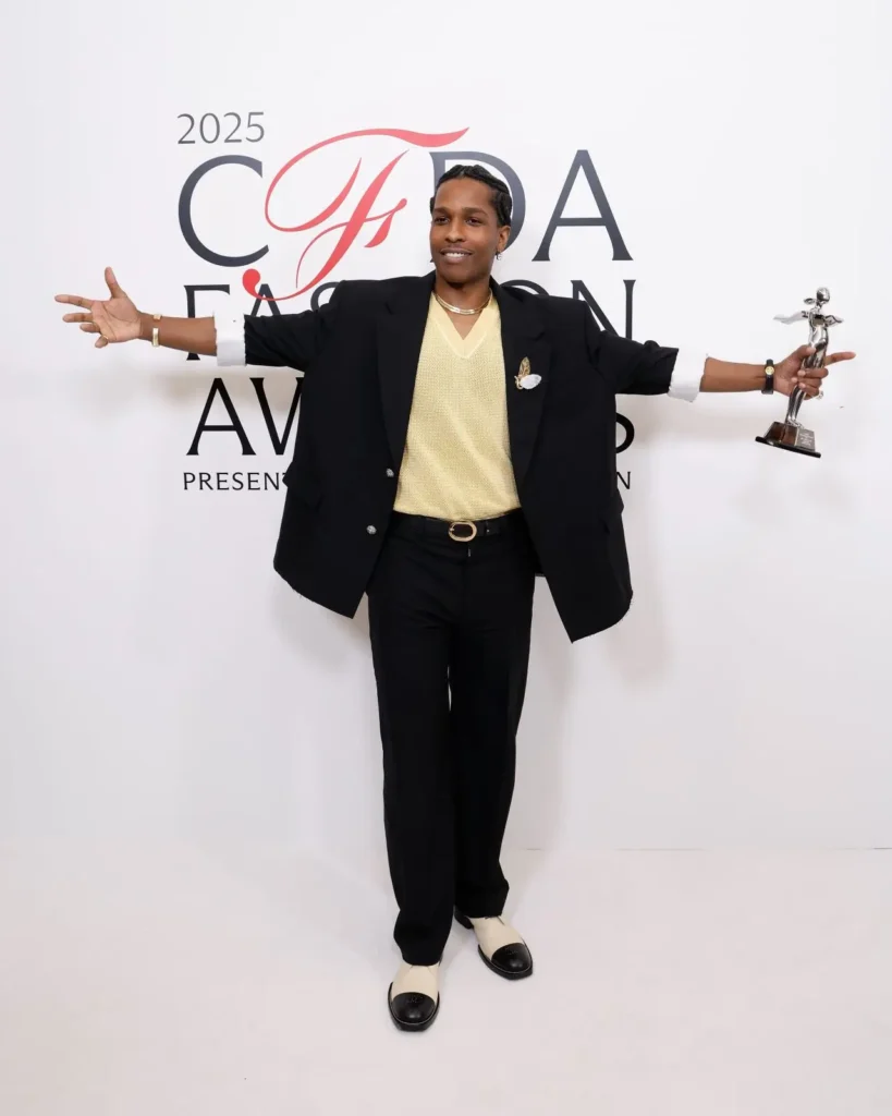 A$AP Rocky poses with his CFDA Fashion Icon Award at the 2025 CFDA Fashion Awards in New York, wearing a black tailored suit over a pale yellow knit shirt, accessorized with layered gold jewelry and two-tone shoes, extending his arms in celebration