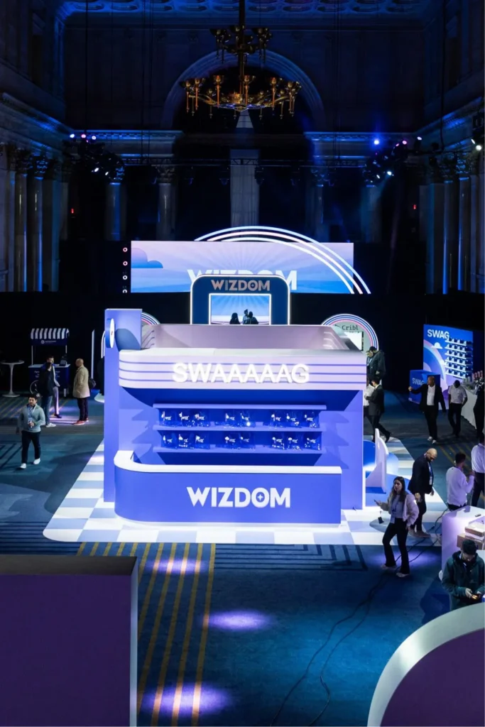 Interior view of the Wizdom 2025 conference at Cipriani Wall Street, featuring a futuristic diner-style installation illuminated in shades of blue and purple. The large “WIZDOM” display structure, labeled “SWAAAAAAG,” serves as a central merch and engagement station surrounded by attendees and event lighting beneath the venue’s ornate ceiling