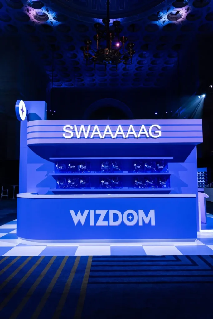 Close-up view of the “WIZDOM” swag station at the Wizdom 2025 event inside Cipriani Wall Street. The installation features bold blue lighting, sleek curved architecture, and illuminated “SWAAAAAAG” signage above transparent shelves filled with branded Wiz gift bags, set against the ornate ceiling of the venue