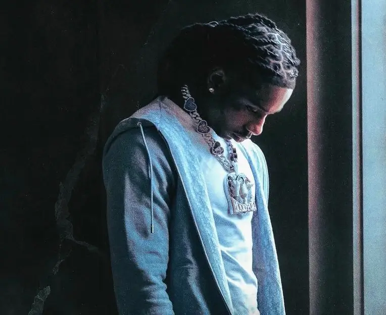 Polo G stands near a window in a dimly lit room, looking down with a reflective, somber expression. He wears a light hoodie over a white shirt and a large diamond chain, while soft blue light highlights his face and braids, creating a moody, introspective atmosphere