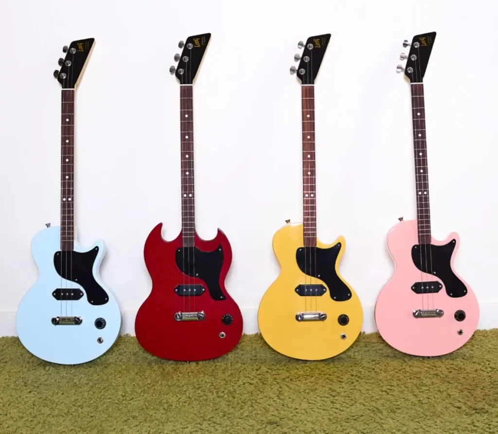 lineup of four Gibson x Loog electric guitars standing upright on green carpet against a white wall, featuring pastel blue, cherry red, TV yellow, and blush pink finishes, each with three strings and black pickguards, showcasing the playful yet classic mini Les Paul and SG-inspired designs