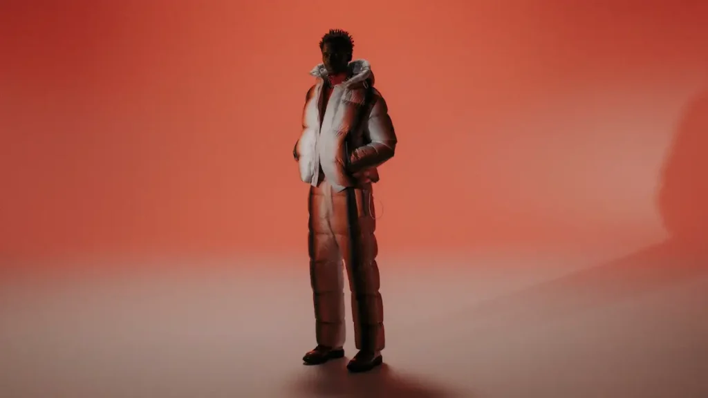 A model stands against a warm orange gradient backdrop wearing a reflective silver and copper puffer jacket with matching quilted trousers from TATRAS’ “Layers of Japanese Craft” collection. The soft lighting and long shadows highlight the sculptural volume and fluid craftsmanship of the outfit